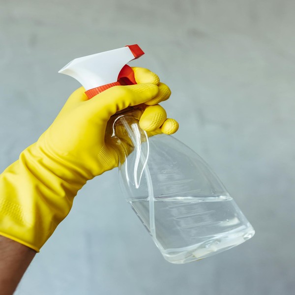 Yellow gloved hand holding a transparent cleaning spray bottle against a neutral background.